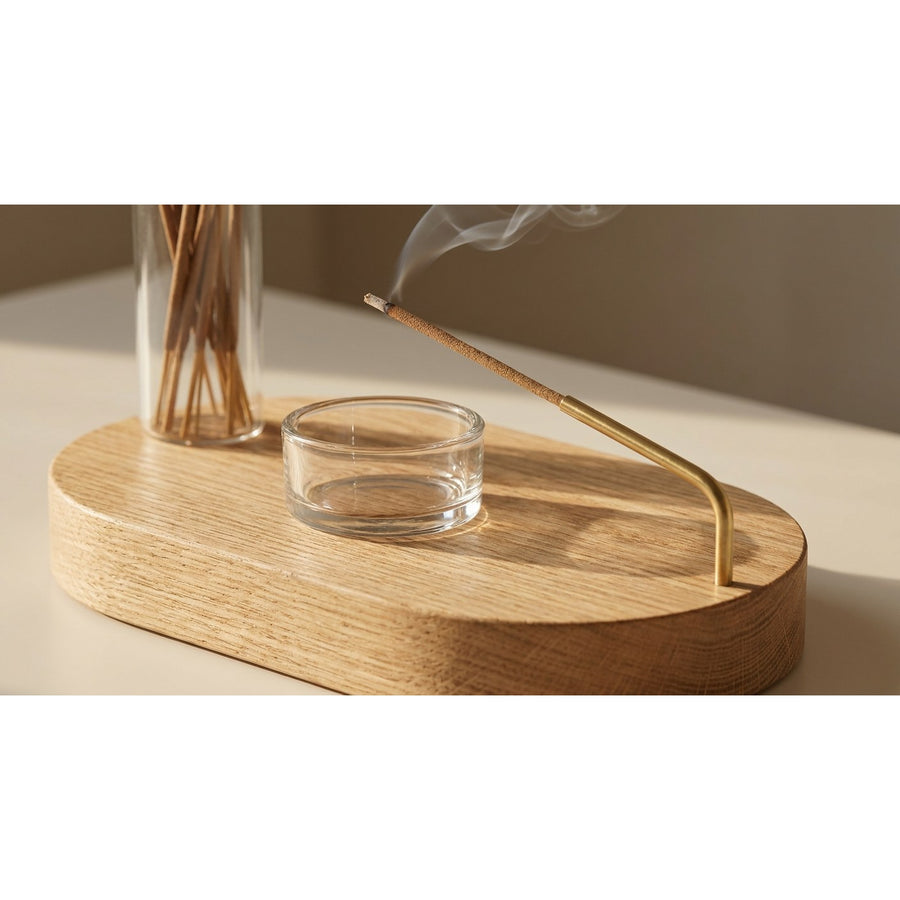 Close-up of burning incense stick in brass holder over glass ash catcher on wooden base with rising smoke