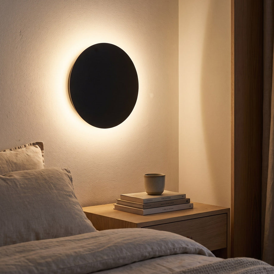 Black round LED wall light used as evening lighting above a nightstand
