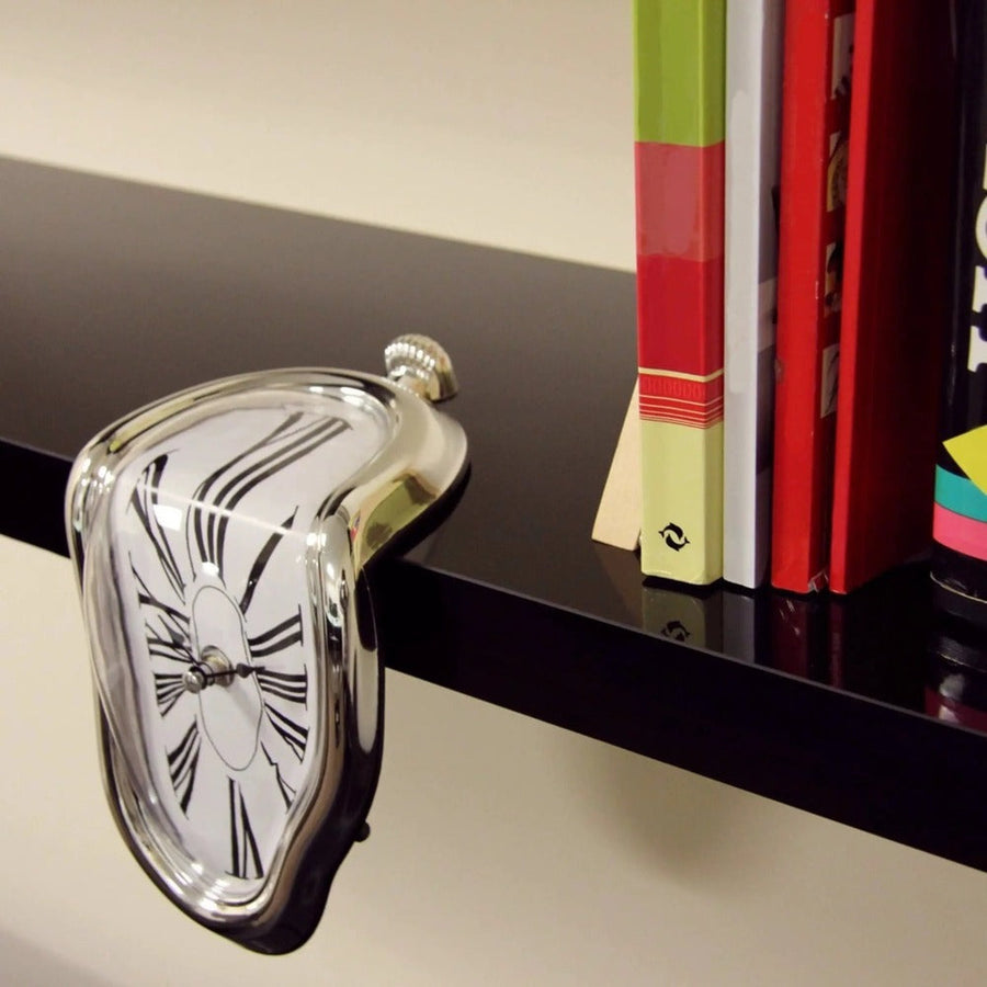 Silver melting wall clock resting among books on a dark shelf.